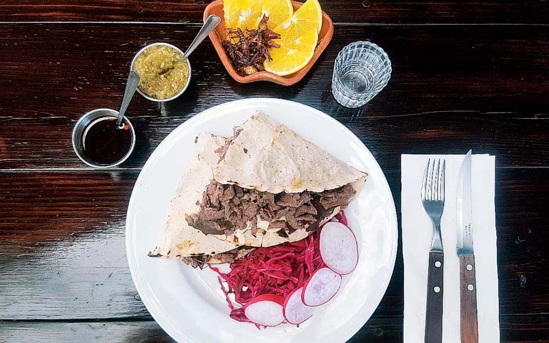 Presentación de tlayuda con varios complementos como salsas, shots de mezcal y chapulines